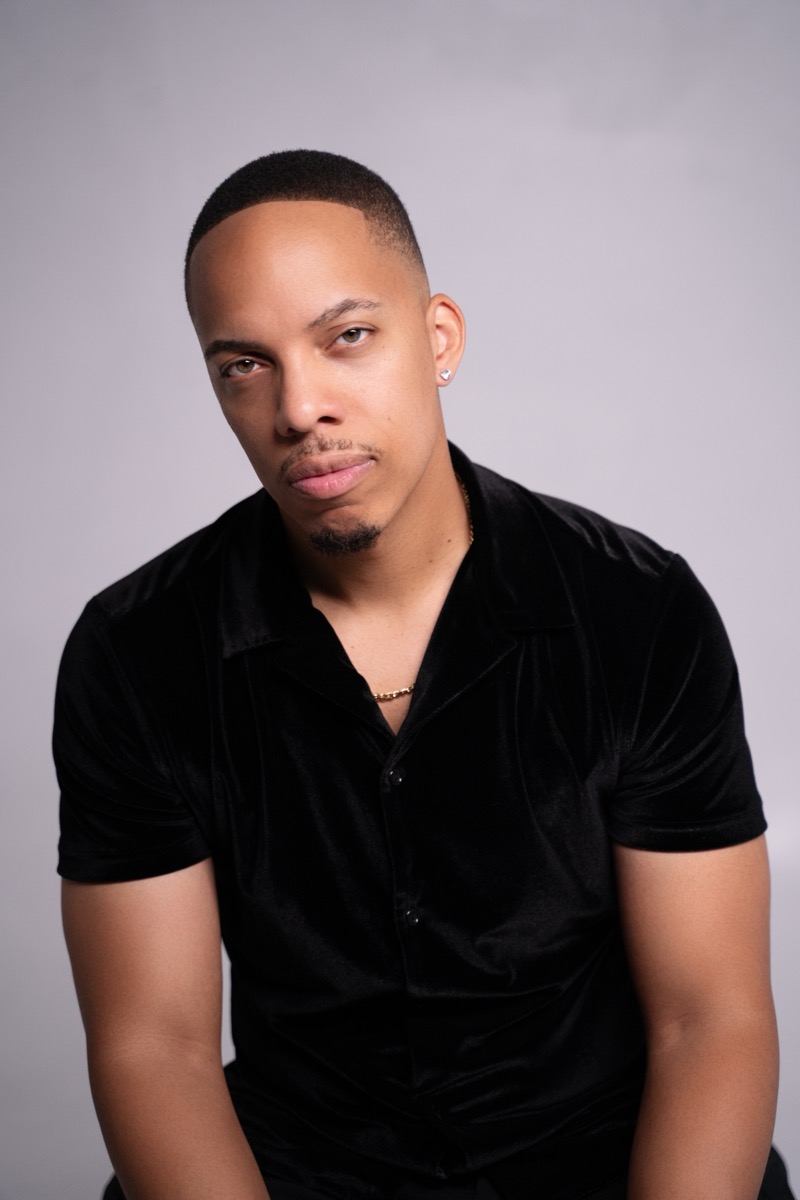 Studio headshot of Jerel Crockett in a black velvet camp-collar shirt, looking straight to camera against a pale grey backdrop.