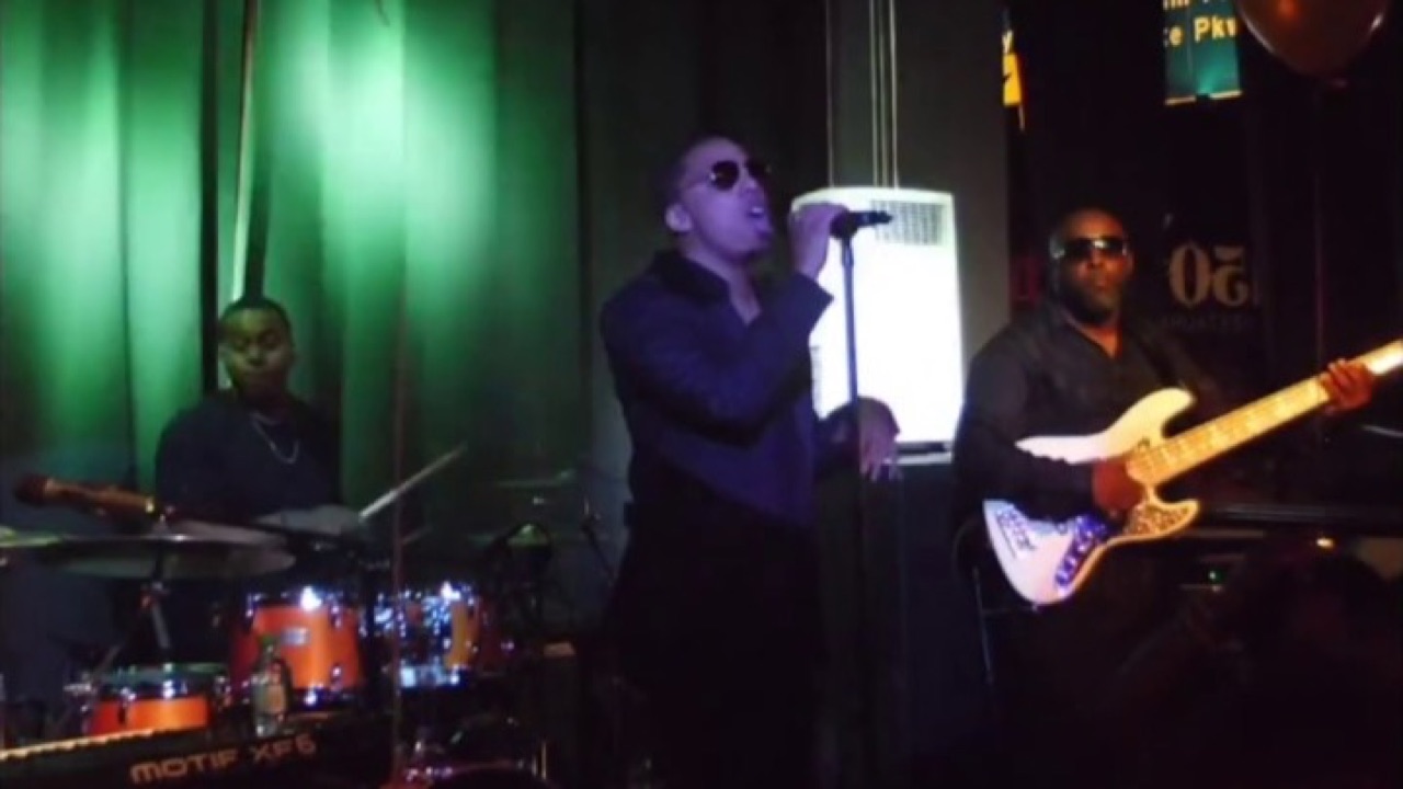 Jerel Crockett center stage on a handheld microphone with his band around him, drummer to one side and bassist to the other, under green stage light in a dark room.